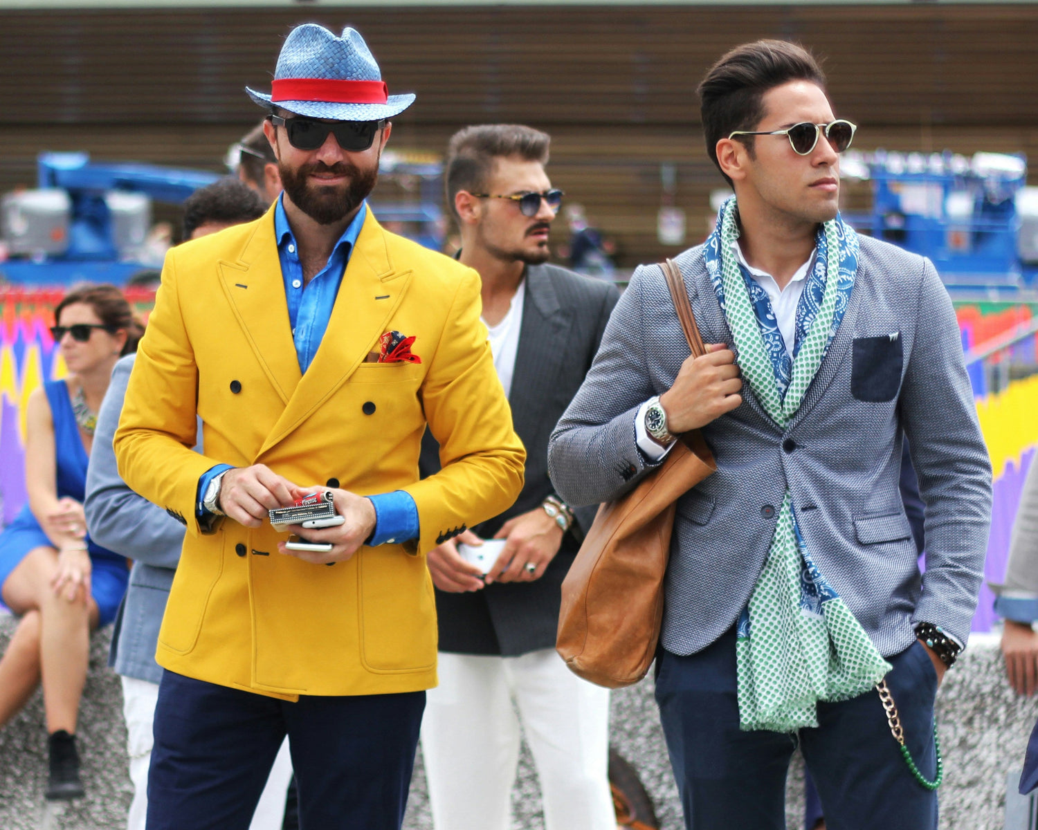 Two men in stylish outfits walking together, one in a yellow jacket and blue hat, the other in a gray blazer with a scarf.